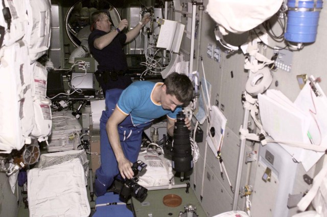 Cosmonaut Sergei K. Krikalev (foreground), flight engineer, works with cameras in the service module (Zvezda) on the International Space Station, while astronaut William M. (Bill) Shepherd, Expedition 1 commander, busily goes about chores in the background. The photograph was taken by cosmonaut Yuri P. Gidzenko, Soyuz commander, using an electronic still camera (ESC). This was one of the first still pictures to be downlinked from the station since the Expedition 1 crew boarded it earlier in the week.