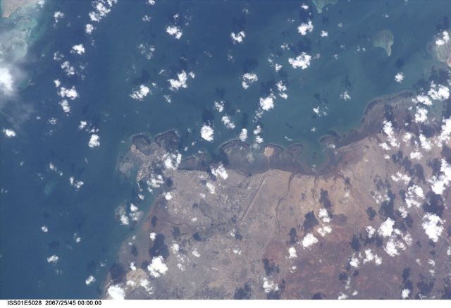 Djibouti, the capital city of the country of Djibouti, can be seen in this northeast-looking view taken from the International Space Station (ISS) with a digital still camera using a 400mm lens. Djibouti, scarcely 100 years old, sits on the western shore of an isthmus in the Gulf of Tadjoura, an arm of the Gulf of Aden. Djibouti is surrounded by a rugged and bleak landscape, that has a dry and hot climate. The population of Djibouti has grown from an estimated 96,000 in 1973 to over 330,000 in 1991 mainly due to the influx of refugees from the neighboring, war torn countries of Ethiopia and Somalia. With its strategically located port, Djibouti’s economic importance results from the large transit trade it enjoys as the terminus of a railroad line from Addis Ababa in Ethiopia. The city has seen an increase in tourism in the past decade due a large number of cruise ships visiting the port. Besides tourism, salt production and shipbuilding and repair are other major industries. Below the center of the image, the long runway of the Djibouti/Ambouli International Airport is visible. Coral reefs are discernible in the upper left and upper right quadrants of the image.