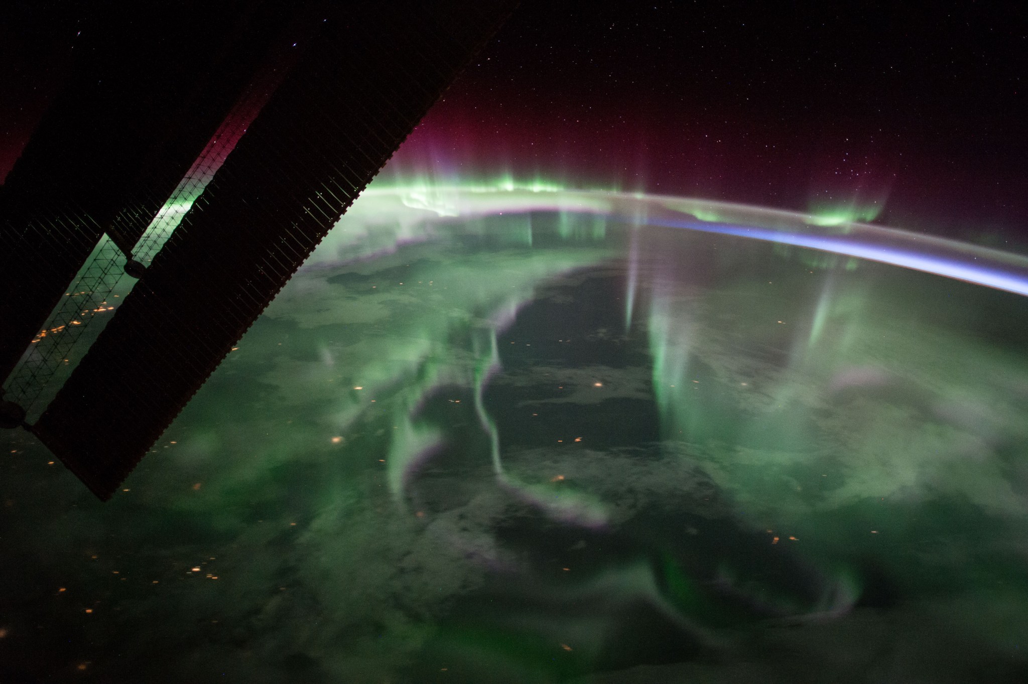A green, fog-like haze stretches over Canada. At the edges of Earth’s atmosphere, the light takes on a purple-red color, eventually fading into the darkness of space. Some stars are faintly visible. At top left, a portion of the solar arrays of the International Space Station is backlit against the planet.