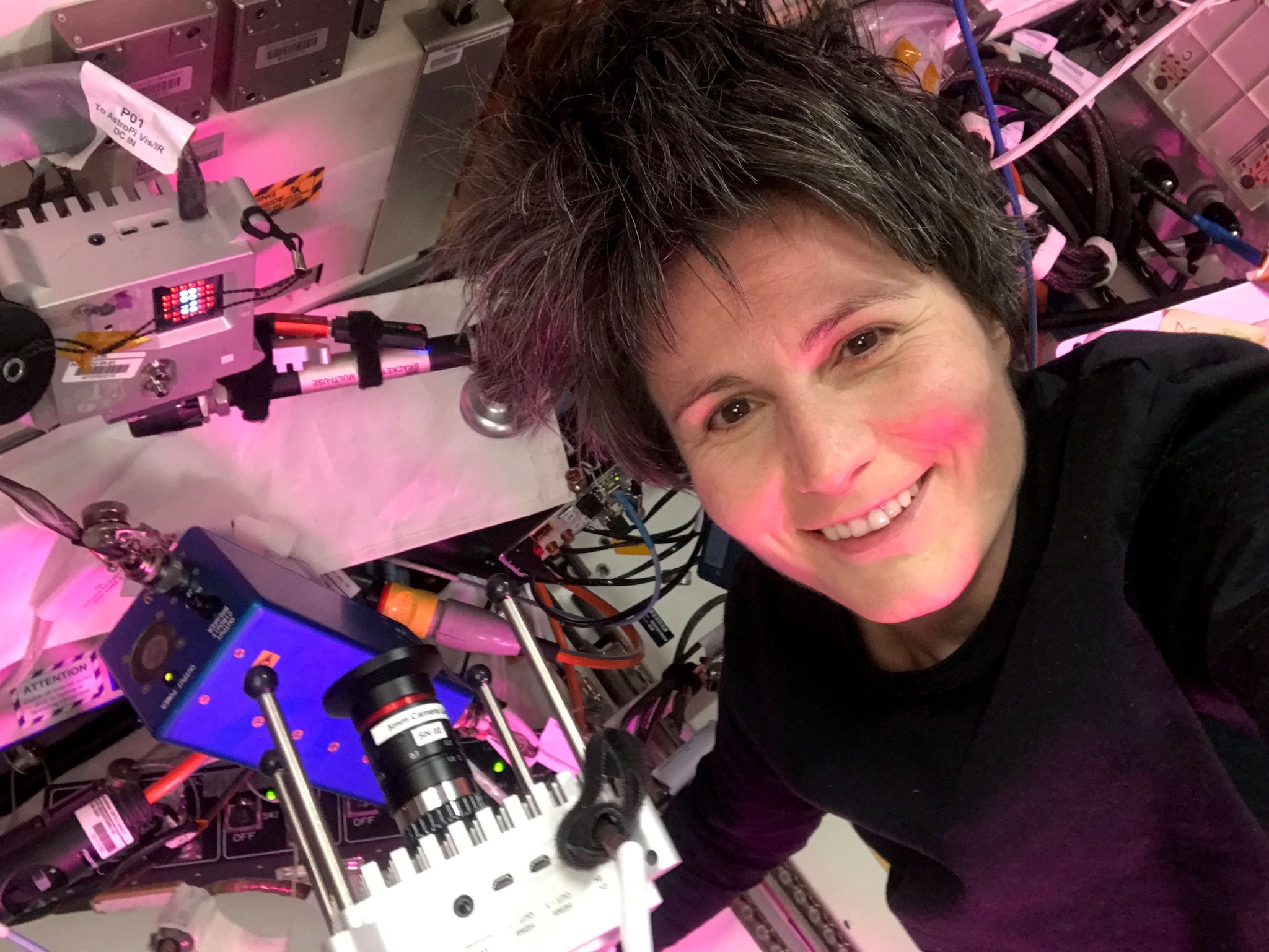 Samantha Cristoforetti, wearing a long-sleeved purple shirt, smiles at the camera. Hardware floating next to her includes a partly visible AstroPi with a black camera attached and a silver AstroPi with red and white lights.