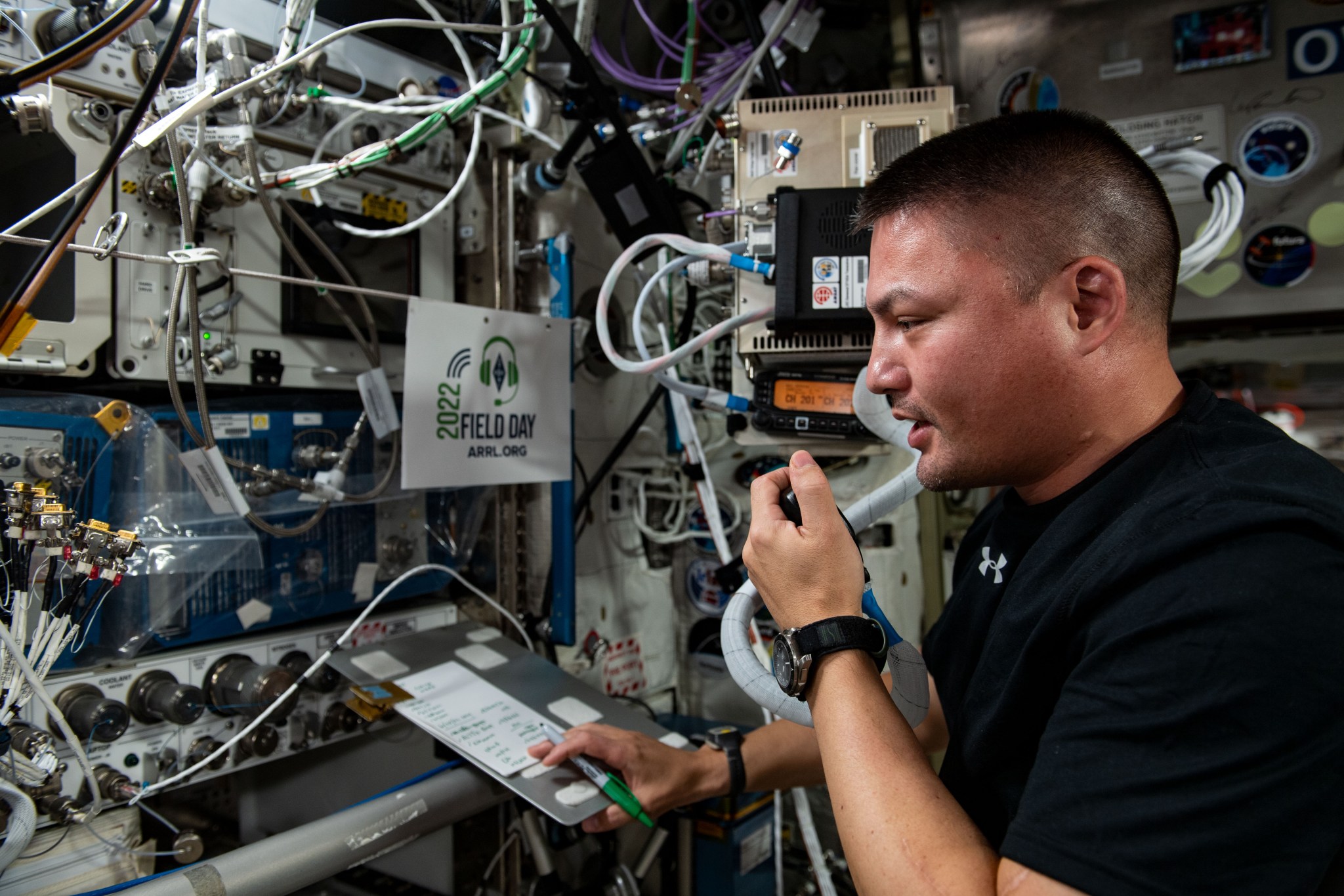 Astronaut Kjell Lindgren, wearing a black shirt, faces the control panel of the station’s ham radio set. He is holding a notepad and pen in his right hand and speaking into the microphone in his left hand.