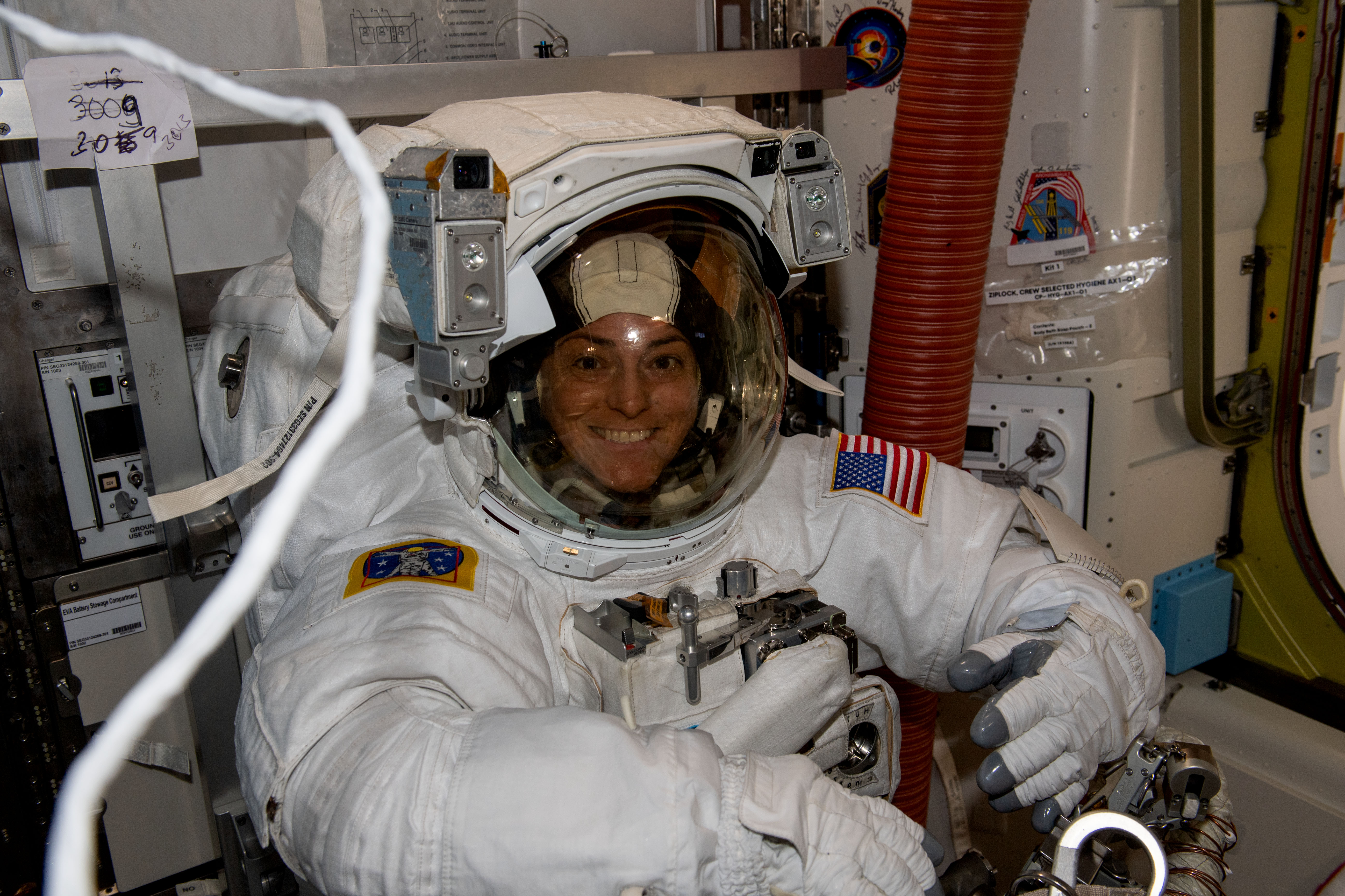 Astronaut Nicole Mann faces the camera and smiles. She is wearing a white spacesuit, which has an American flag patch on the left arm, and another patch on the right arm.