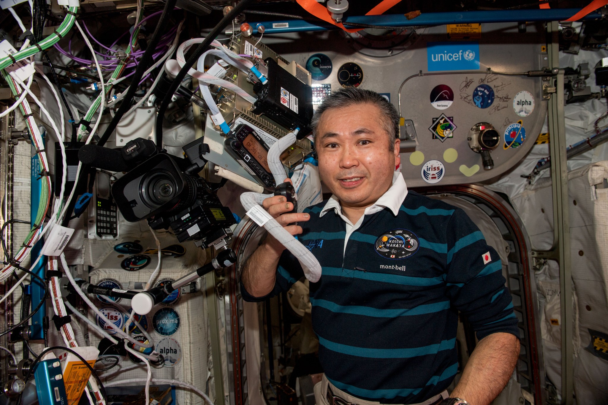 Koichi Wakata, wearing a striped shirt, faces the camera, holding the ham radio mic in his right hand. It is attached by a large grey cable to the radio set above his head. A large camera and tangles of wires are next to him.
