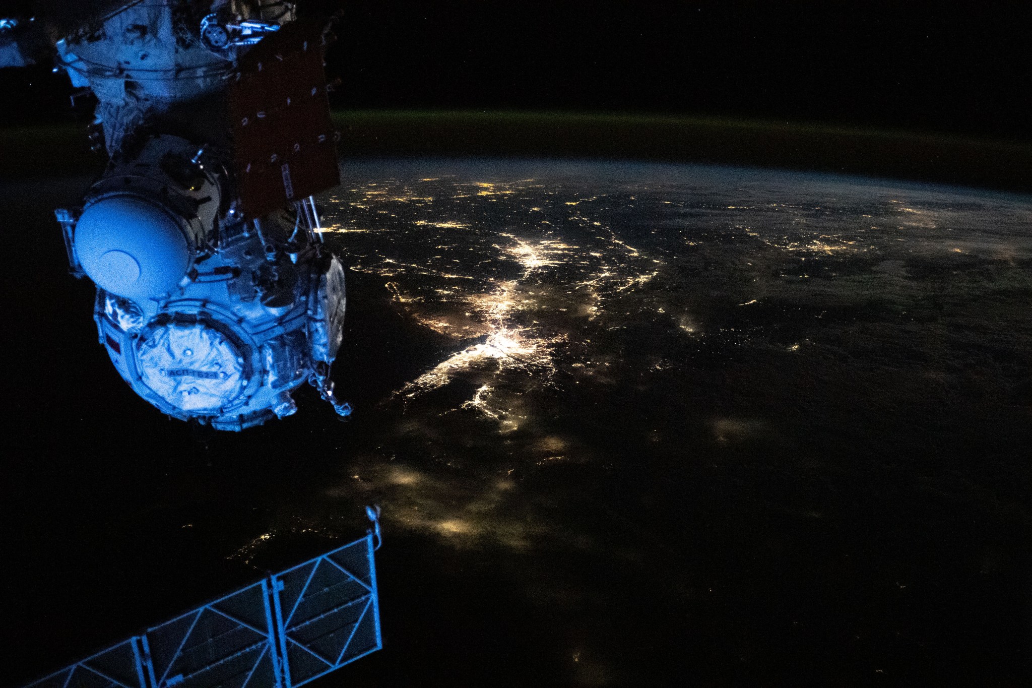 The city lights of the northeastern United States are visible from the International Space Station. The rest of the surrounding land is mostly dark, as is space. At left, part of the orbital lab and a docking port on a Russian space station module are visible.