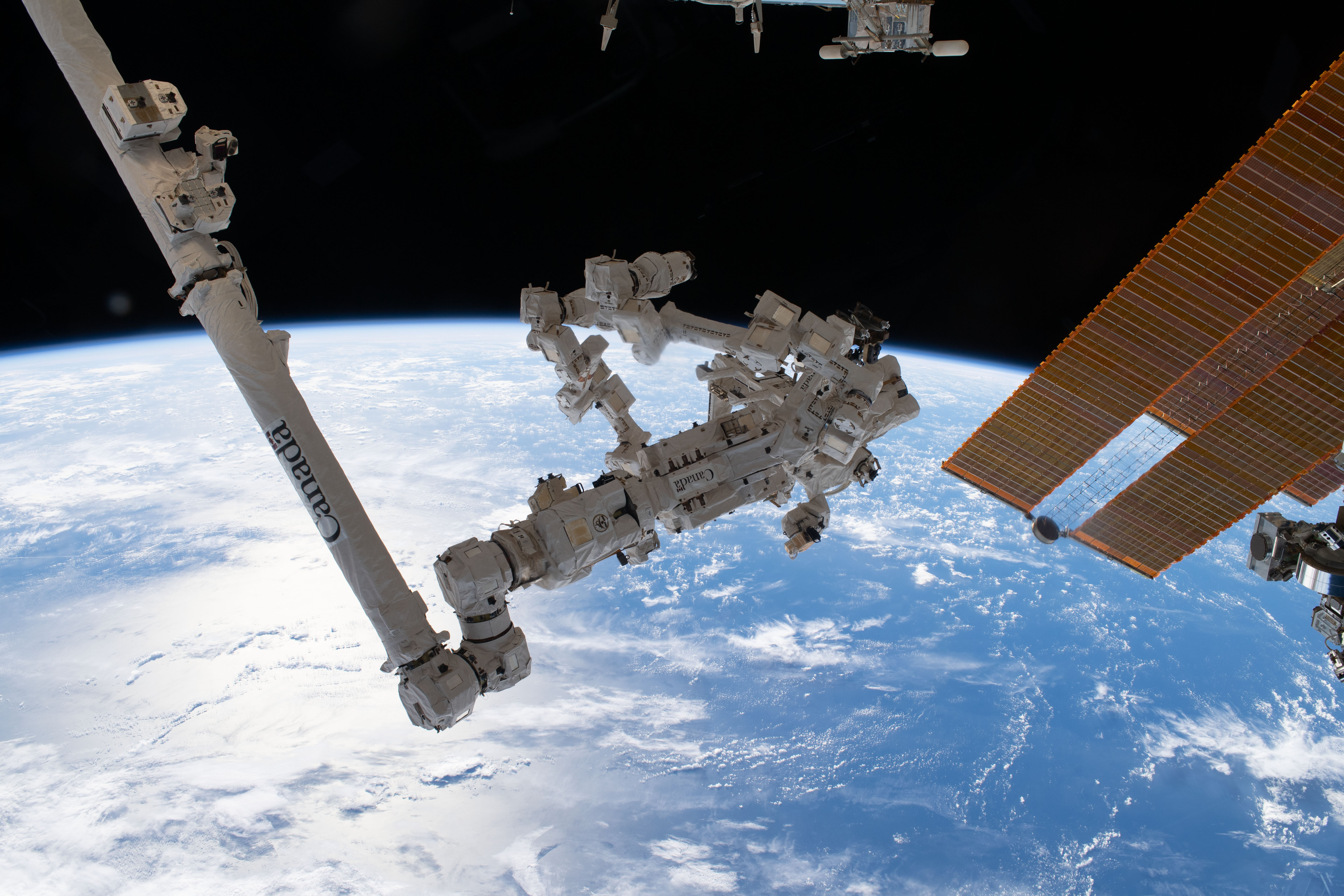 The sun's glint beams off a clear blue north Atlantic Ocean as the International Space Station orbited 261 miles above. In the foreground, a pair of main solar arrays drape across the right with the Canadarm2 robotic arm and its fine-tuned robotic hand Dextre extending from the left.