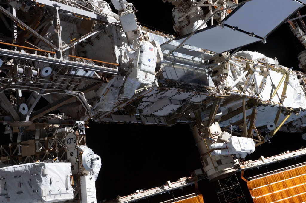 Expedition 70 Flight Engineers Loral O'Hara (center) and Jasmin Moghbeli (lower right), both from NASA, are pictured tethered to the International Space Station's port truss structure during a spacewalk to replace one of the 12 trundle bearing assemblies on the port solar alpha rotary joint, which allows the arrays to track the Sun and generate electricity to power the station.
