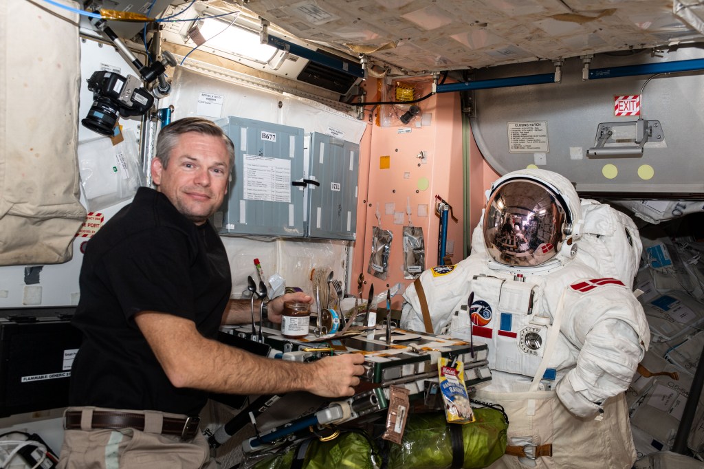 ESA (European Space Agency) astronaut and Expedition 70 Commander Andreas Mogensen poses for a playful portrait with an empty spacesuit across from him inside the galley in the International Space Station's Unity module.