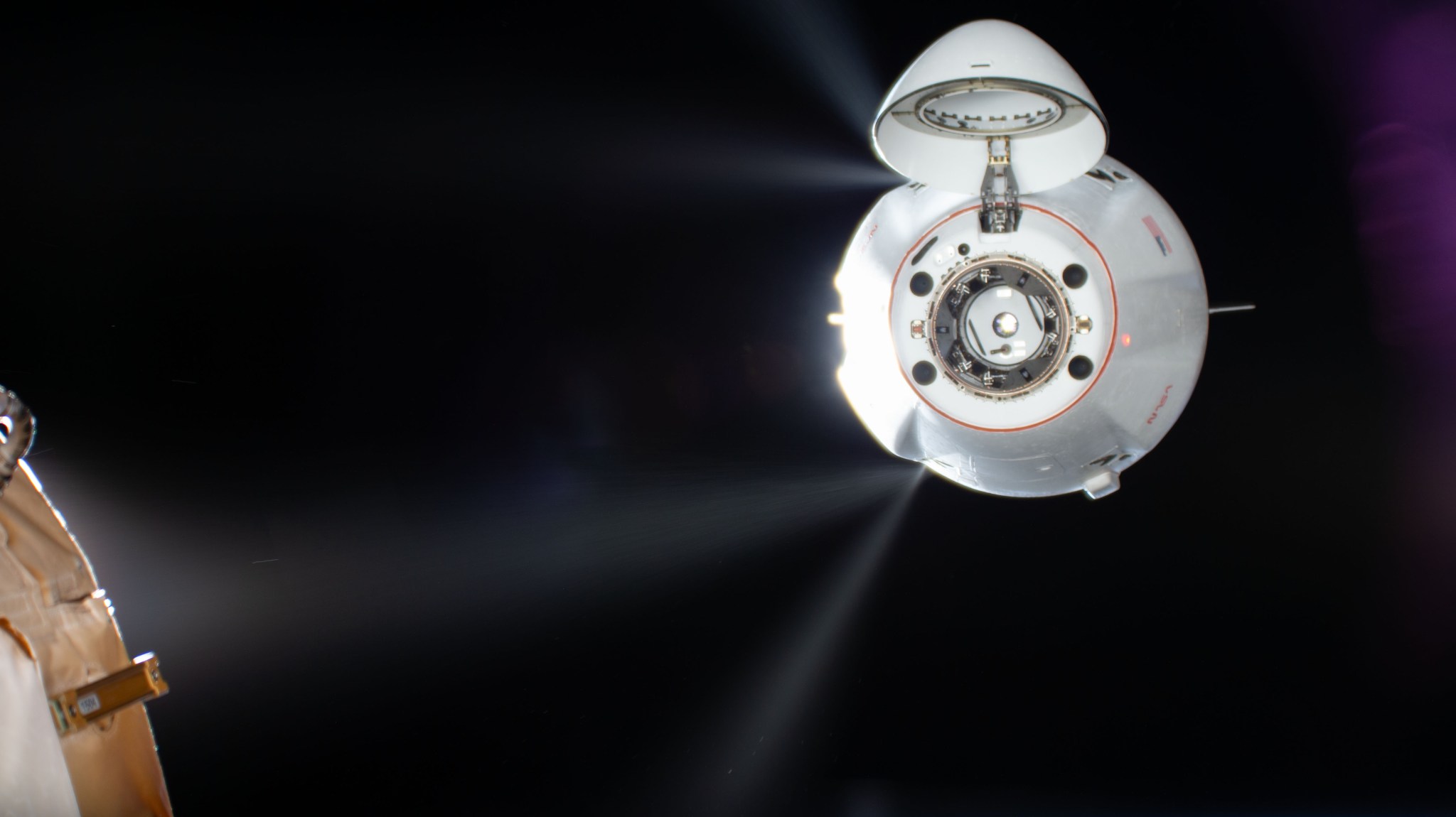 A white Dragon spacecraft approaches the station against the blackness of space. Its top hatch is open, revealing the docking ring, and jets of propulsion fuel are visible shooting from its top and bottom on the left side. A portion of the station is visible at the bottom left of the image.