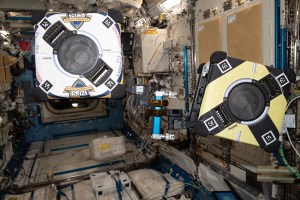 Two Astrobee free-flying robotic assistants are pictured during an experiment demonstrating how the toaster-sized robotic devices can use a small robotic arm and grasp handrails to maneuver throughout the space station.