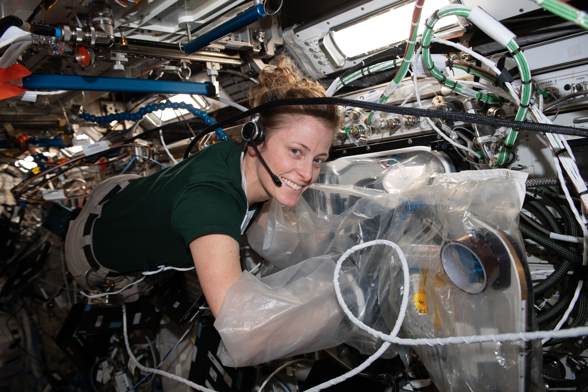O’Hara is floating horizontally, smiling at the camera, her arms inside plastic sleeves of the BioFabrication Facility, which is connected to the wall to her right. She is wearing a green t-shirt and khaki pants and has a headset on. Several cords float in front of her.