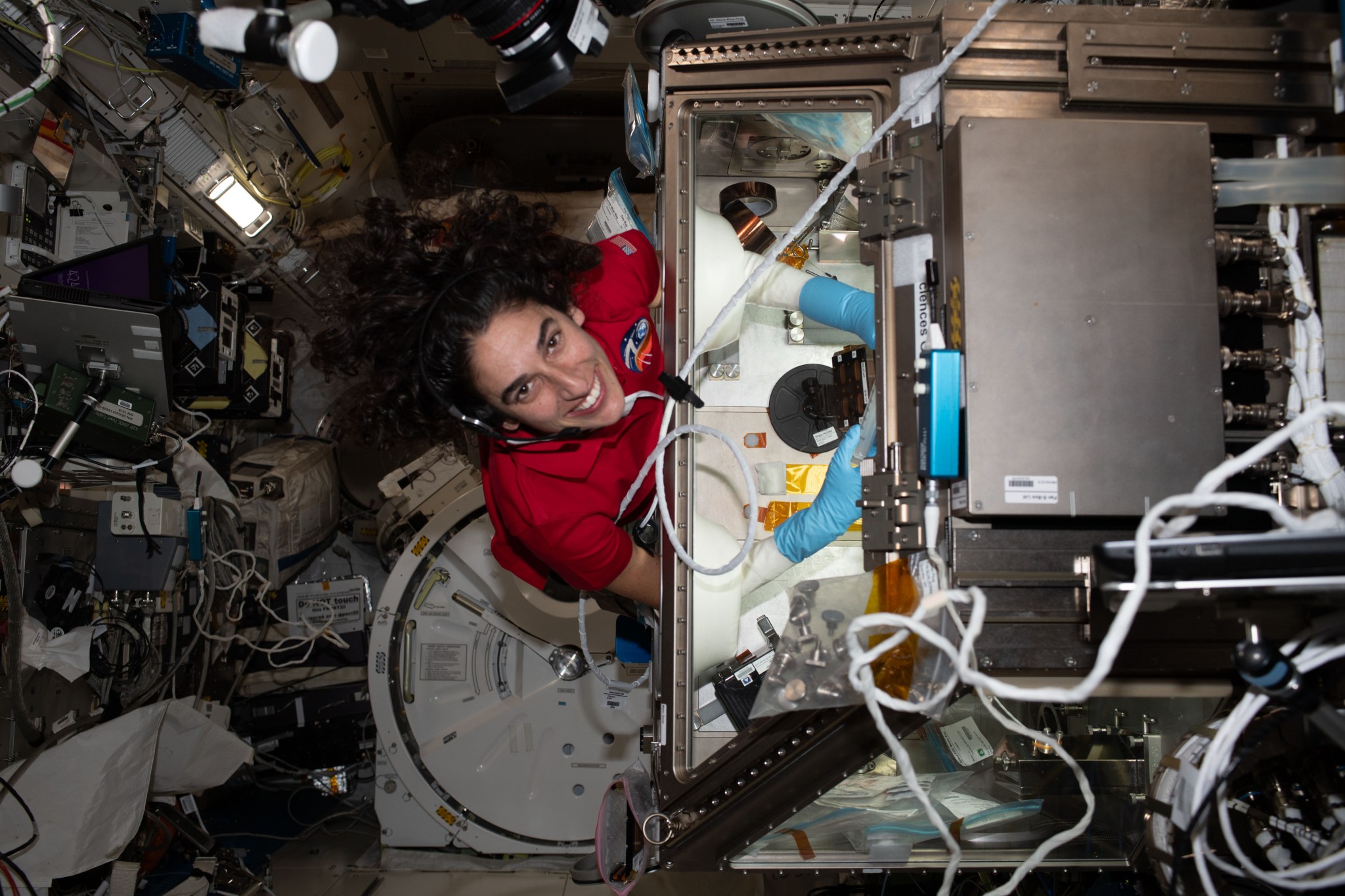 Jasmine Moghbeli, wearing a red polo shirt and a headset, looks up and smiles at the camera. Her arms are inside a large, clear glovebox used to contain experiments. Equipment, laptops, cords, and lights cover the walls behind her.