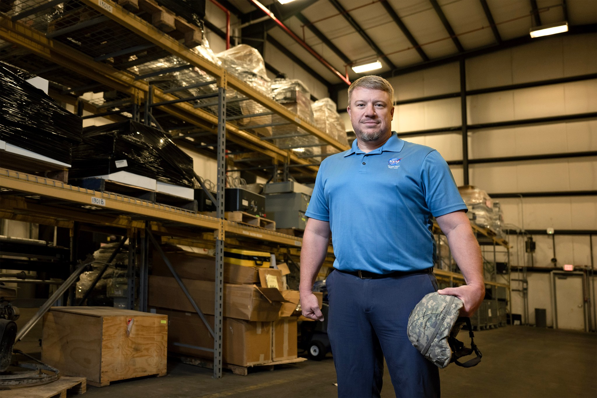 A white man in a blue NASA shirt stands in a warehouse looking building.