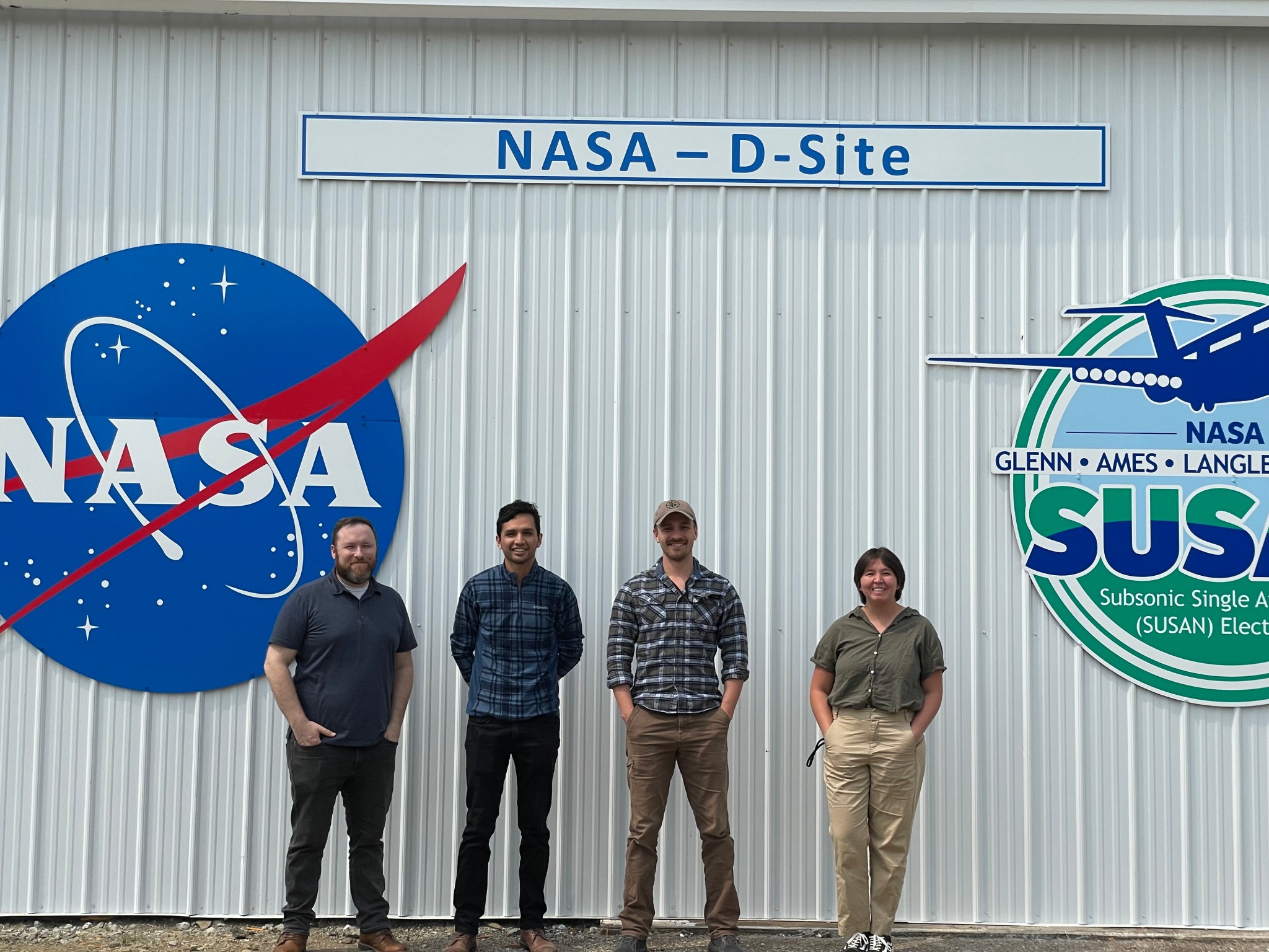 : A group of four students pose in front of a white metal building. On the side of the building is a NASA meatball logo, a sign that says, “NASA – D-site,” and a colorful SUSAN aircraft logo.