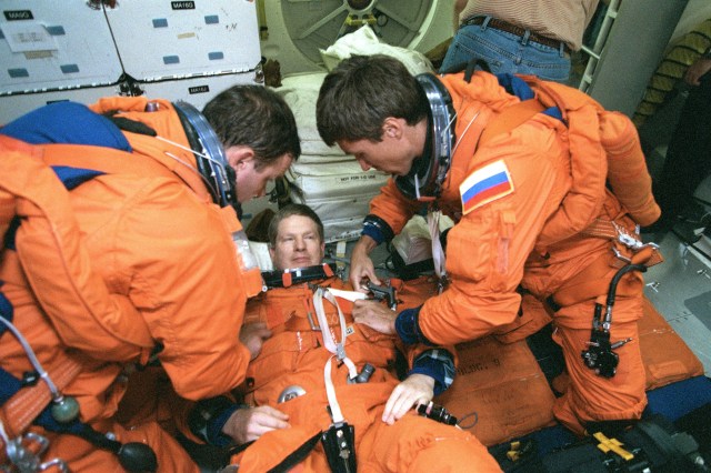 Astronaut William Shepherd, Expedition 1 mission commander, looks on while his two Expedition 1 crewmates apply final touches to his full pressure entry suit as he lies on a couch on the mid deck of a Johnson Space Center trainer. Cosmonaut Yuri Gidzenko, left, is Soyuz commander, and cosmonaut Sergei Krikalev is flight engineer. Scheduled to come back from their ISS stay aboard the Space Shuttle Discovery, the three were participating in a rehearsal of their duties during shuttle descent.