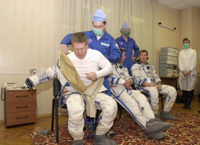 Expedition 1 commander William M. (Bill) Shepherd gets help with his Sokol suit prior to a simulation at Baikonur. Also donning suits are Soyuz pilot Yuri P. Gidzenko (seated at center) and flight engineer Sergei K. Krikalev.