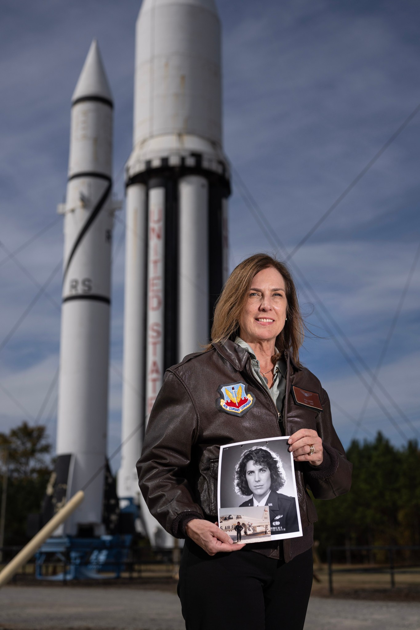 A woman in a leather bomber jacket holds a photo of herself in the military