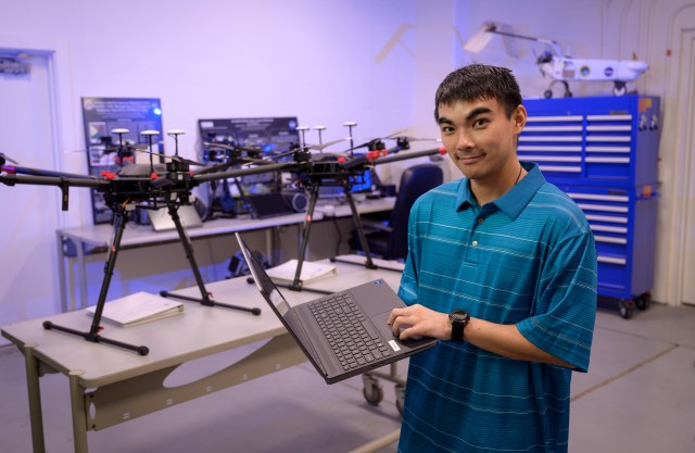 Evan Kawamura holds a laptop beside a table holding two drone aerial vehicles.
