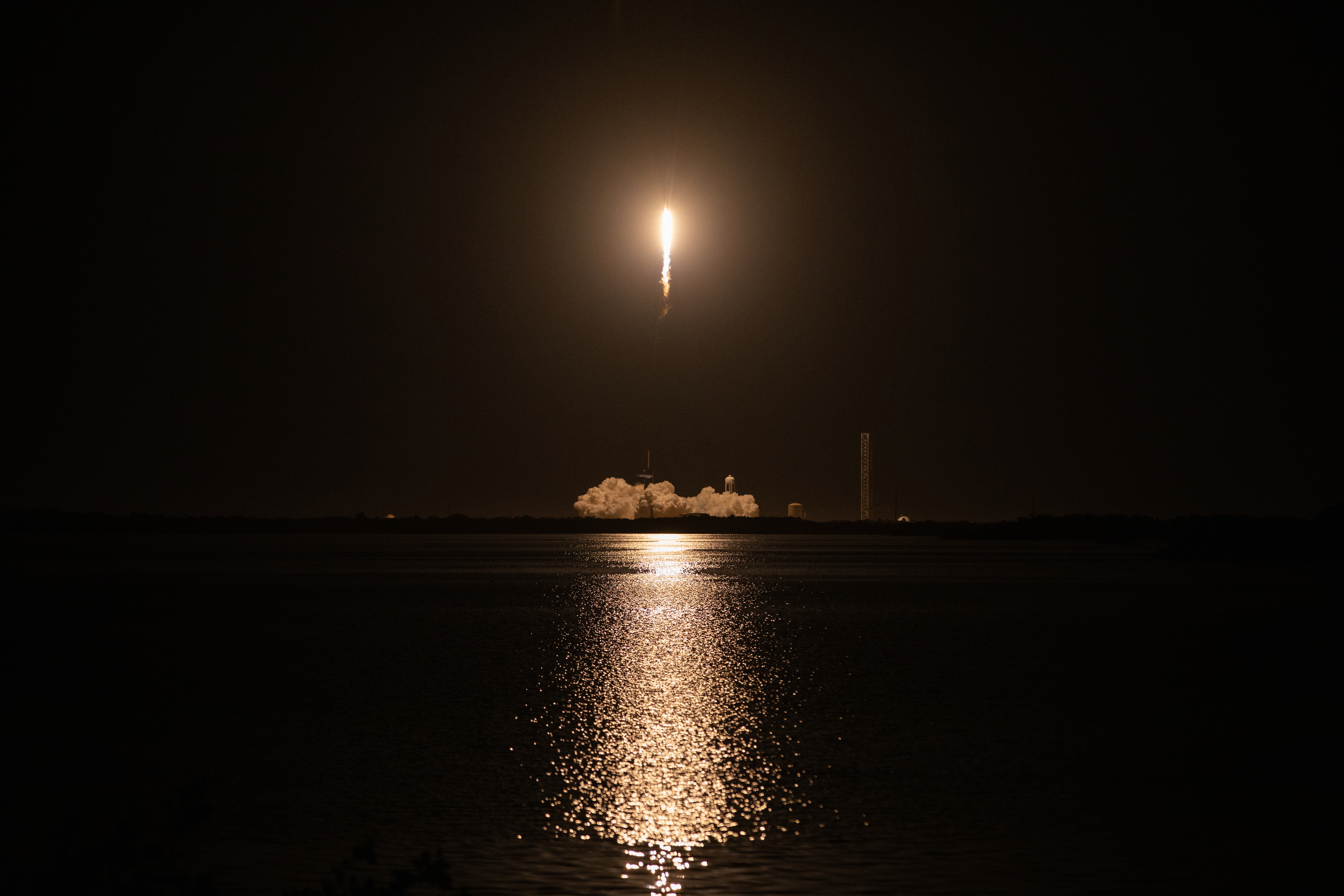 SpaceX's Cargo Dragon launches from Launch Complex 39A at NASA's Kennedy Space Center in Florida in its 29th commercial resupply mission. The rocket's engines are glowing brightly as it takes off through the dark night sky, and the light illuminates the water below.