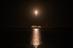 SpaceX's Cargo Dragon launches from Launch Complex 39A at NASA's Kennedy Space Center in Florida in its 29th commercial resupply mission. The rocket's engines are glowing brightly as it takes off through the dark night sky, and the light illuminates the water below.