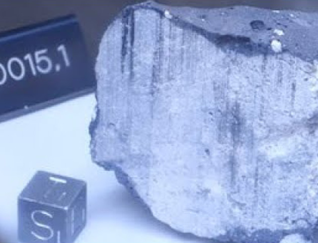 Close-up view of a rock sample returned from the Moon during the Apollo missions, with a small cube next to it for size reference and a sample marker with the number 60015.1 visible