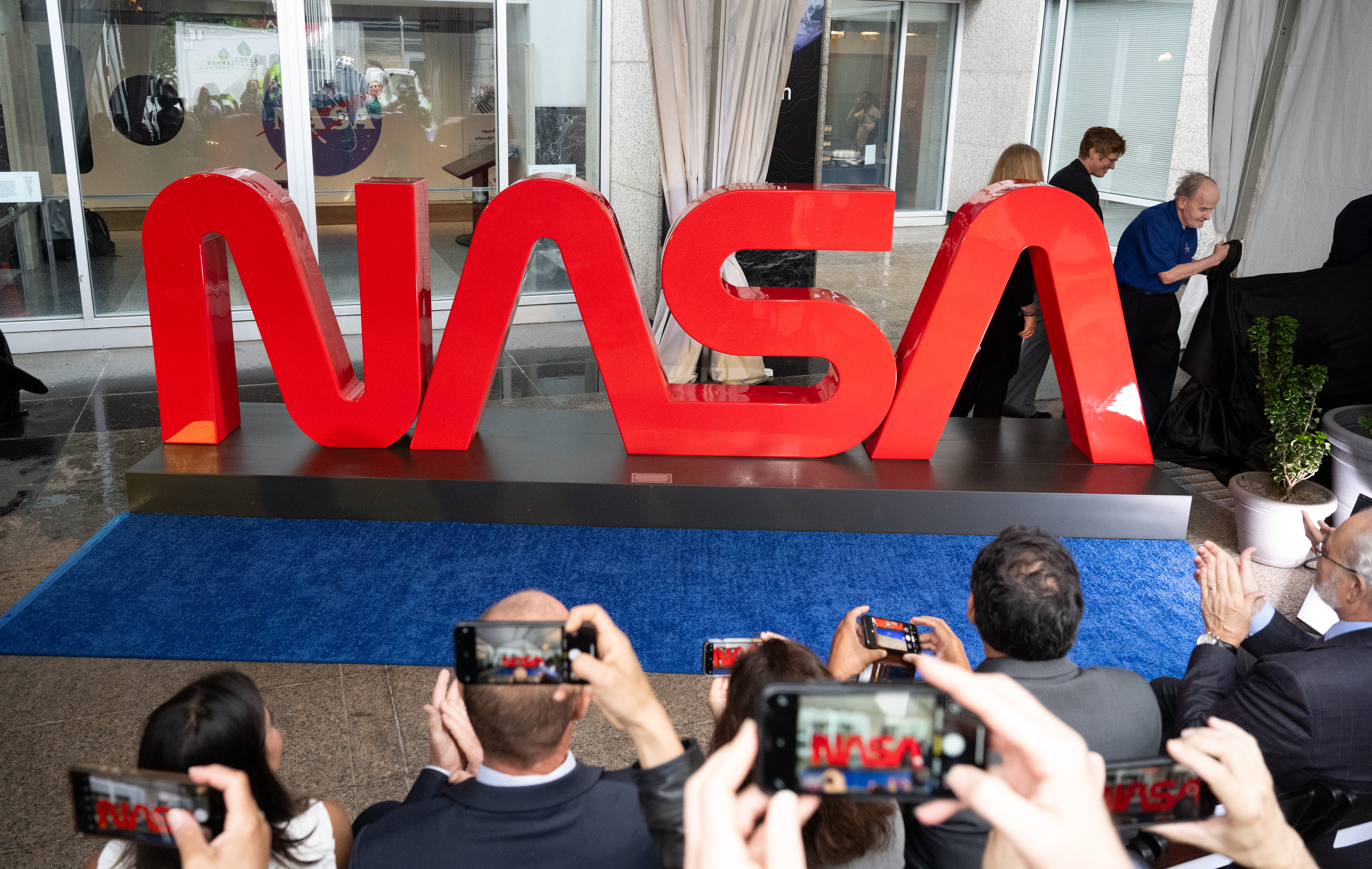 A red sign reading "NASA" in the "worm" logotype created in the 1970s stands before a crowd of people clapping and taking pictures. The red three-dimensional letters rest on a black platform, which is on a blue carpet in front of the Earth Information Center at NASA Headquarters in Washington.