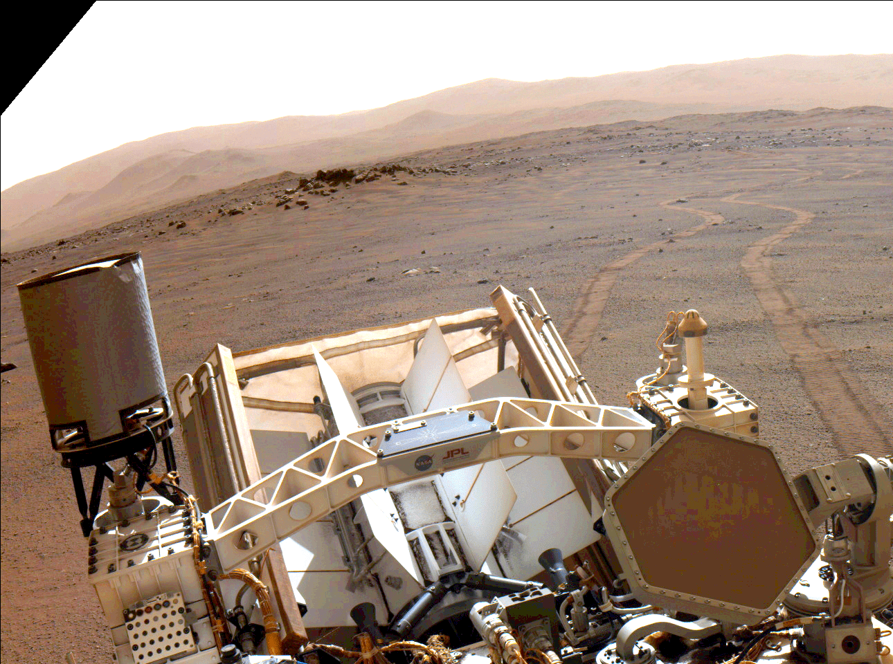 A close-up view of the Multi-Mission Radioisotope Power System on the back end of the Perseverance rover. The rover’s tracks snake through the Martian sand toward a clay-colored mountain range.
