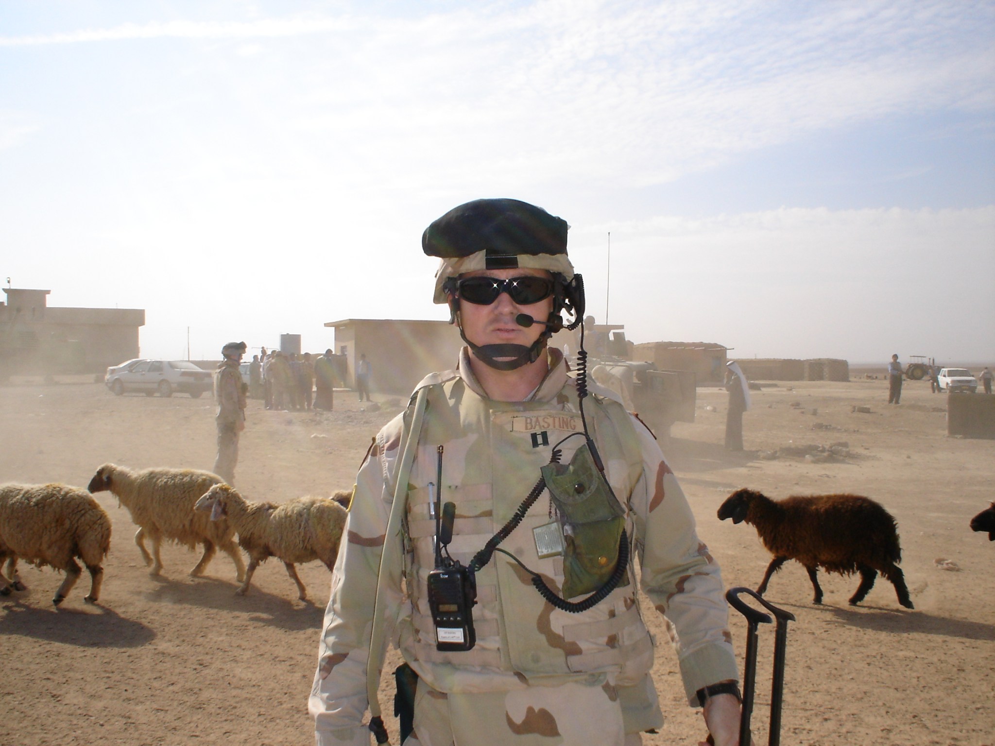 A man in a soldier uniform stands in the desert with sheep behind him.