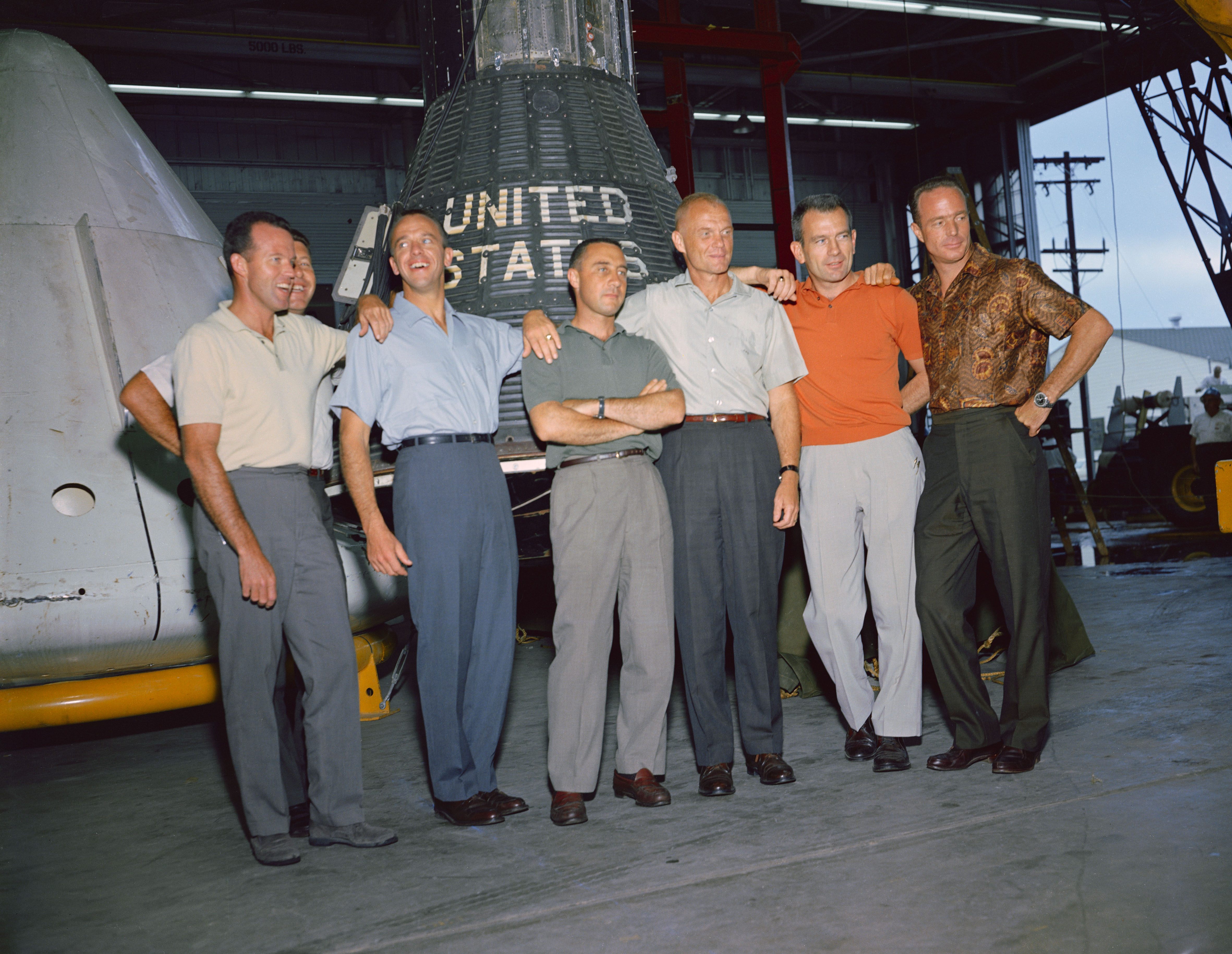 Image of The Mercury 7 astronauts in a more relaxed setting in front of a Mercury capsule at Ellington Air Force Base facilities leased by the Manned Spacecraft Center, now NASA’s Johnson Space Center in Houston