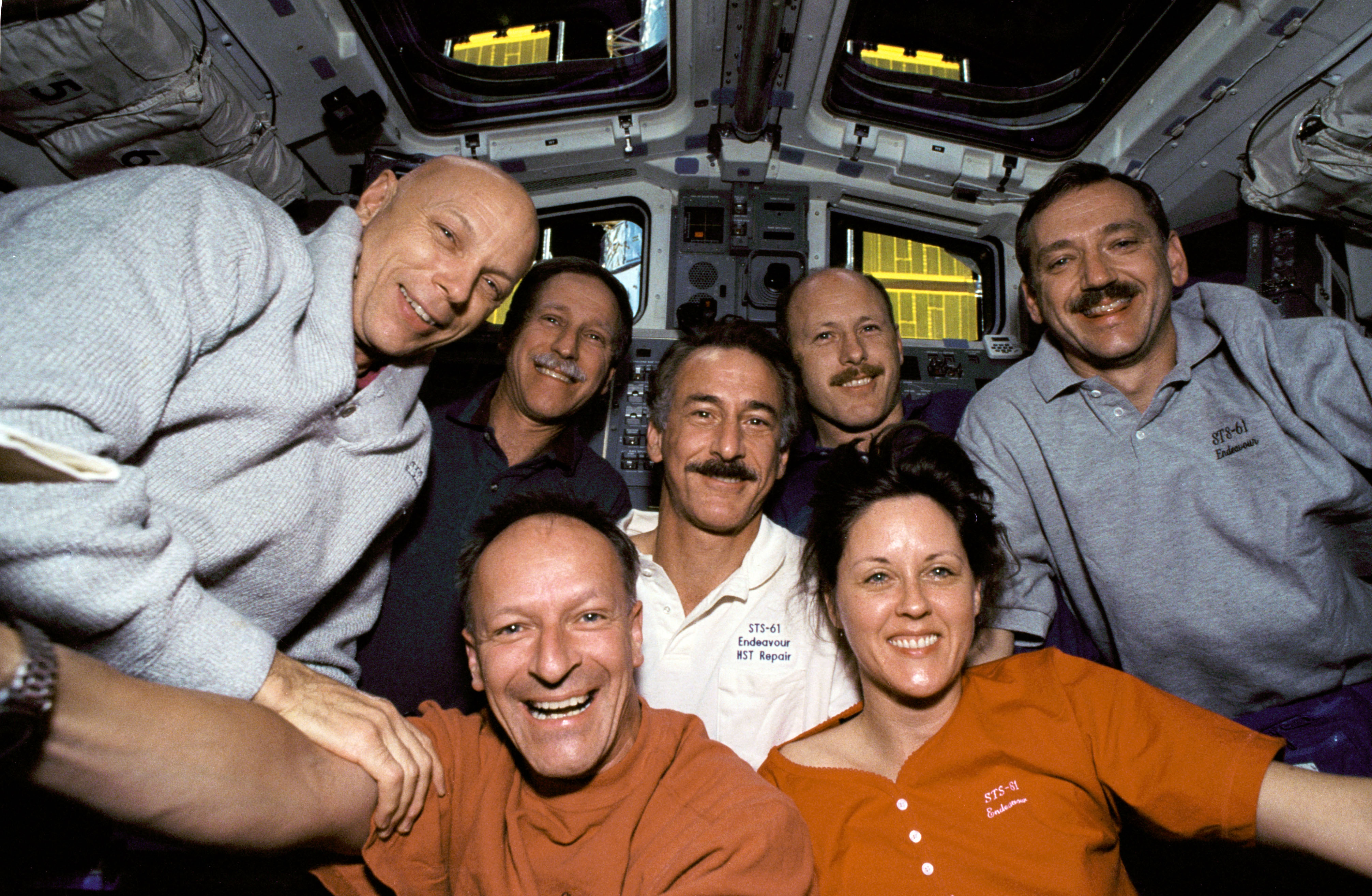 The STS-61 crew poses on Endeavour’s flight deck, with Hubble visible through the windows
