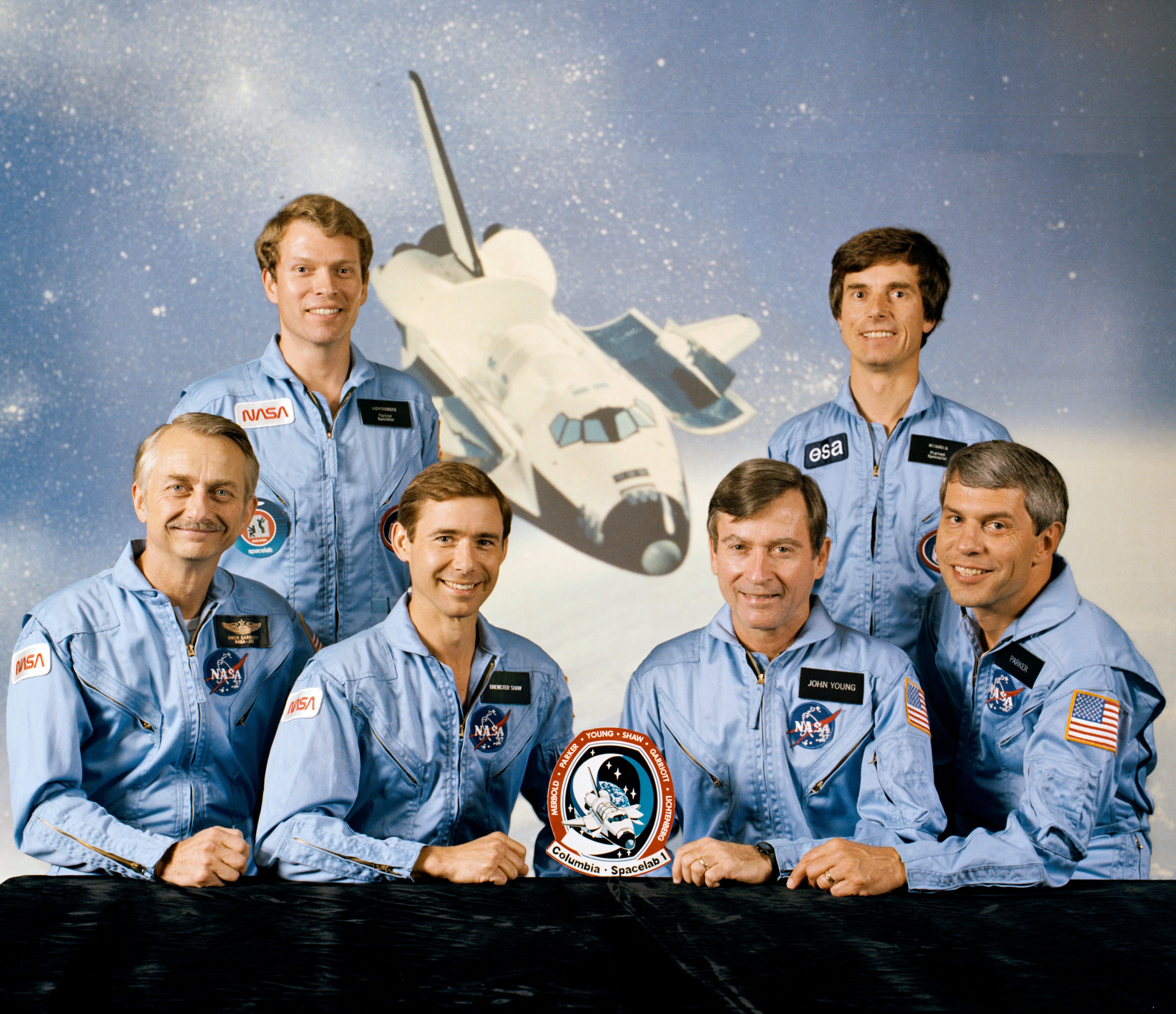 Official photo of the STS-9 crew of Owen K. Garriott, seated left, Brewster H. Shaw, John W. Young, and Robert A.R. Parker; Byron K. Lichtenberg, standing left, and Ulf Merbold of West Germany representing the European Space Agency