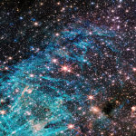 A crowded region of space, full of stars and colorful clouds, more than twice as wide as it is tall. A funnel-shaped region of space appears darker than its surroundings with fewer stars. It is wider at the top edge of the image, narrowing towards the bottom. Toward the narrow end of this dark region a small clump of red and white appears to shoot out streamers upward and left. A large, bright cyan-colored area surrounds the lower portion of the funnel-shaped dark area, forming a rough U shape. The cyan-colored area has needle-like, linear structures and becomes more diffuse in the center of the image. The right side of the image is dominated by clouds of orange and red, with a purple haze.