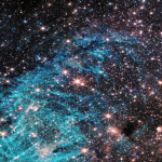 A crowded region of space, full of stars and colorful clouds, more than twice as wide as it is tall. A funnel-shaped region of space appears darker than its surroundings with fewer stars. It is wider at the top edge of the image, narrowing towards the bottom. Toward the narrow end of this dark region a small clump of red and white appears to shoot out streamers upward and left. A large, bright cyan-colored area surrounds the lower portion of the funnel-shaped dark area, forming a rough U shape. The cyan-colored area has needle-like, linear structures and becomes more diffuse in the center of the image. The right side of the image is dominated by clouds of orange and red, with a purple haze.