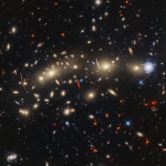 A field of galaxies on the black background of space. In the middle, stretching from left to right, is a collection of dozens of yellowish spiral and elliptical galaxies that form a foreground galaxy cluster. Among them are distorted linear features, which mostly appear to follow invisible concentric circles curving around the center of the image.