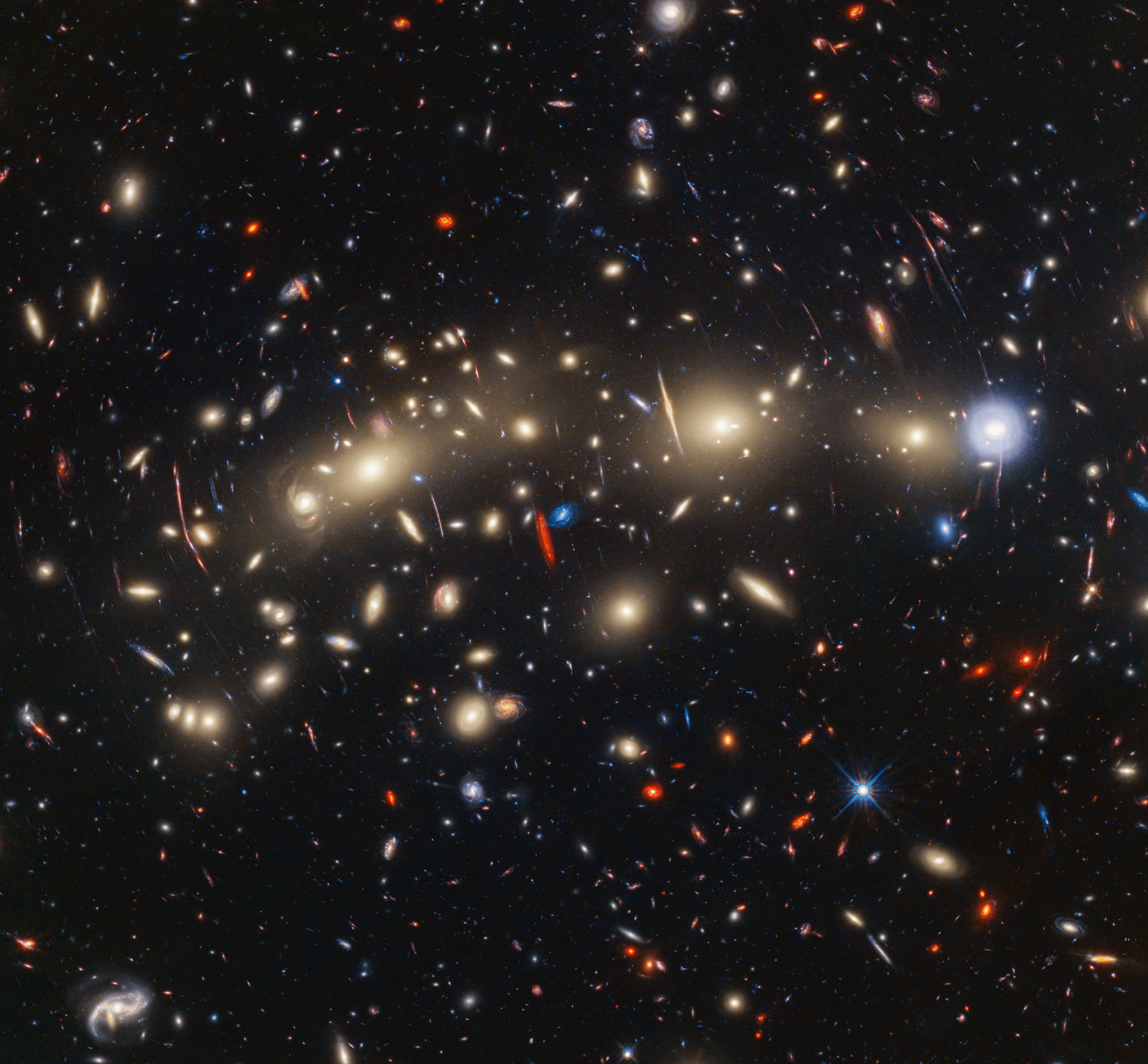 A field of galaxies on the black background of space. In the middle, stretching from left to right, is a collection of dozens of yellowish spiral and elliptical galaxies that form a foreground galaxy cluster. Among them are distorted linear features, which mostly appear to follow invisible concentric circles curving around the center of the image.