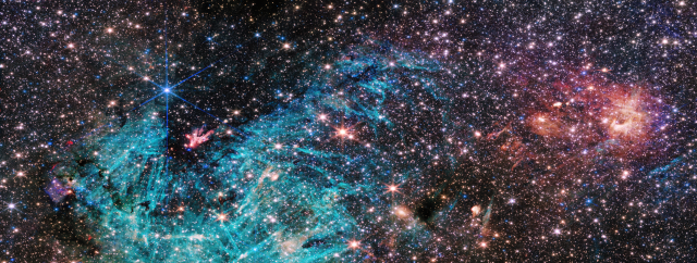 A crowded region of space, full of stars and colorful clouds, more than twice as wide as it is tall. A funnel-shaped region of space appears darker than its surroundings with fewer stars. It is wider at the top edge of the image, narrowing towards the bottom. Toward the narrow end of this dark region a small clump of red and white appears to shoot out streamers upward and left. A large, bright cyan-colored area surrounds the lower portion of the funnel-shaped dark area, forming a rough U shape. The cyan-colored area has needle-like, linear structures and becomes more diffuse in the center of the image. The right side of the image is dominated by clouds of orange and red, with a purple haze.