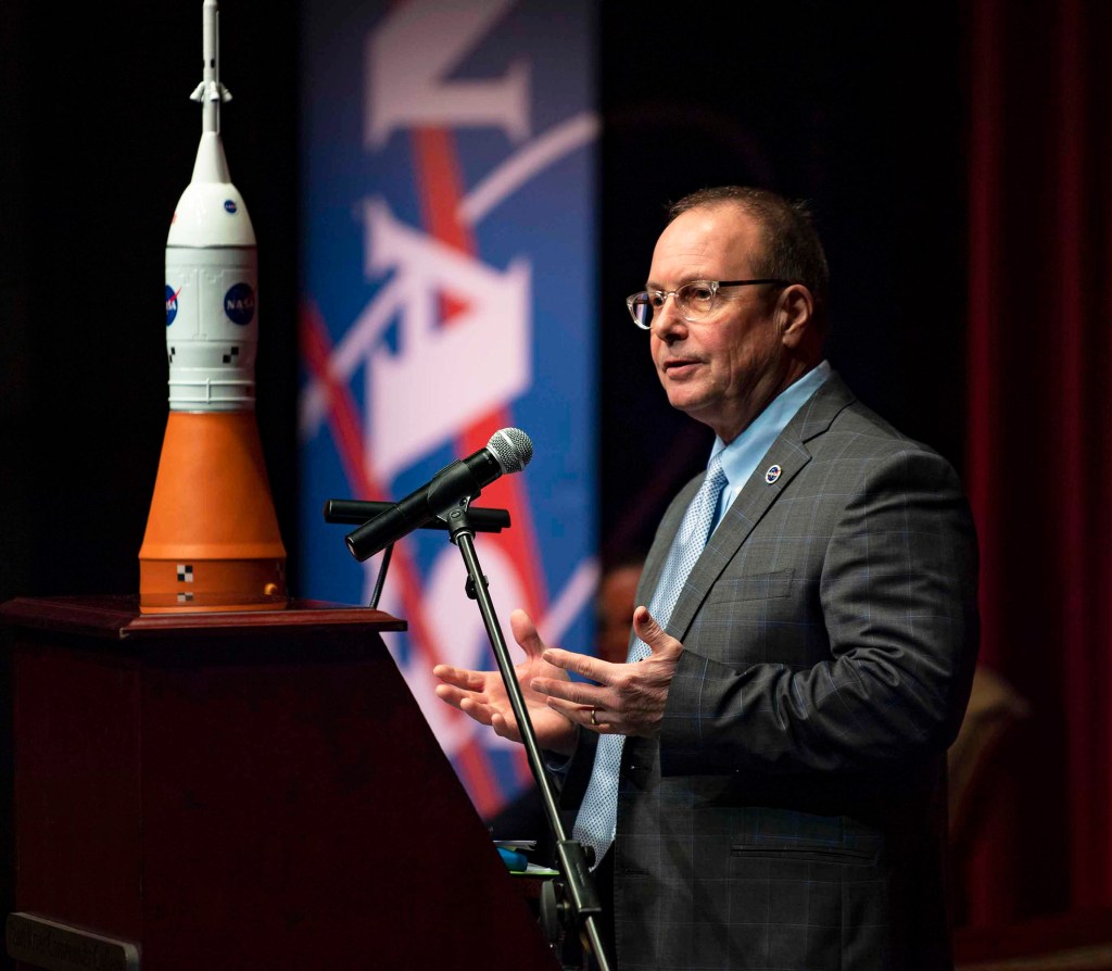 a man wearing in a gray suits stands to speak at podium