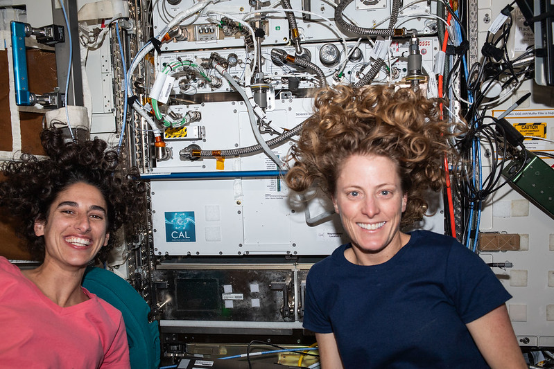 A la izquierda, la astronauta de la NASA Jasmin Moghbeli viste una camisa color rosa, mientras que, a la derecha, la astronauta de la NASA Loral O’Hara viste una camisa azul. Entre ellas hay una vista del Laboratorio de Átomos Fríos en la pared del módulo. Ambas tienen el cabello suelto, flotando por encima de sus cabezas.