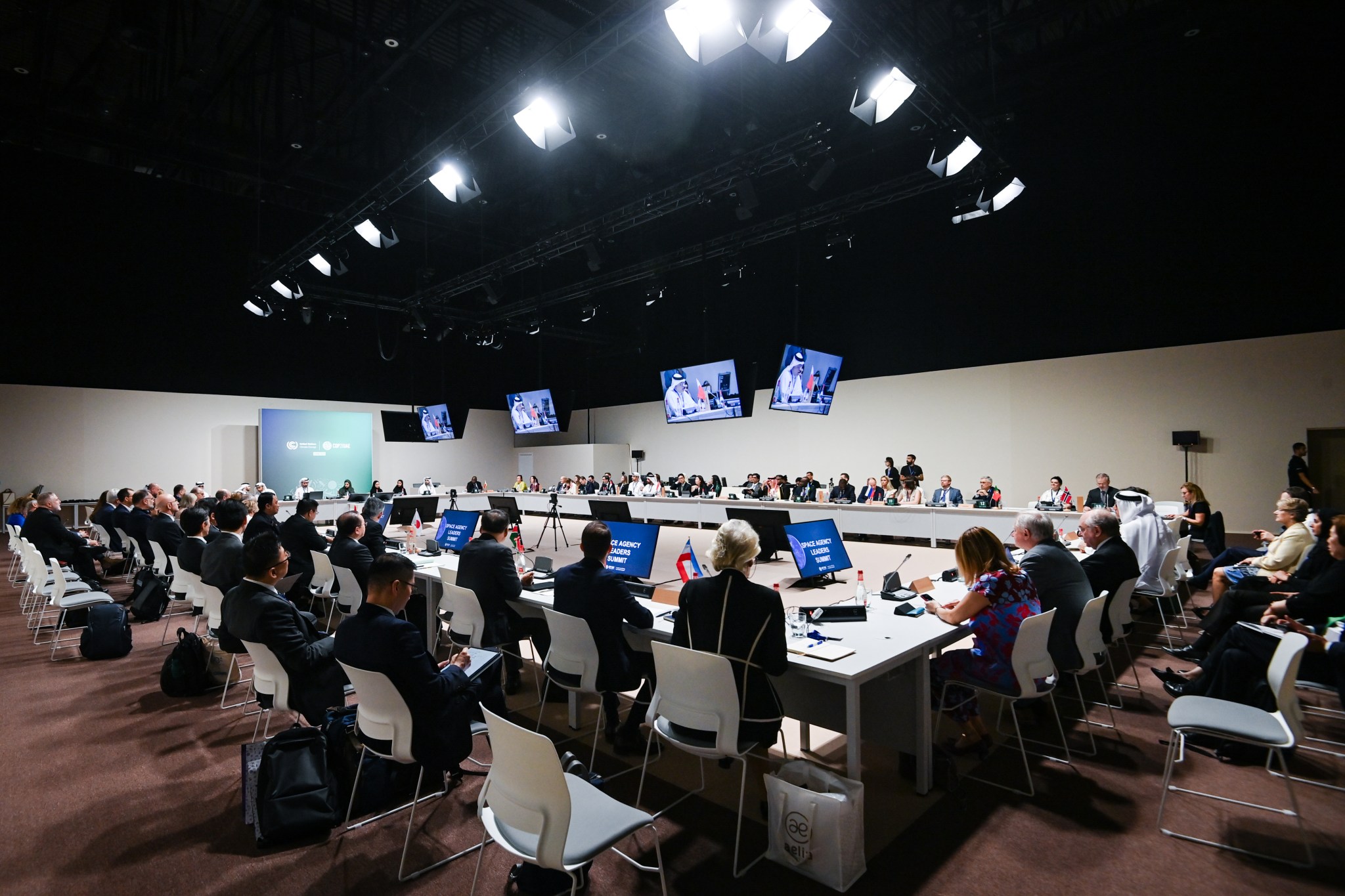 Leaders from two dozen space agencies sit in rows of chairs around a large open rectangular table illuminated under a truss of white lights.