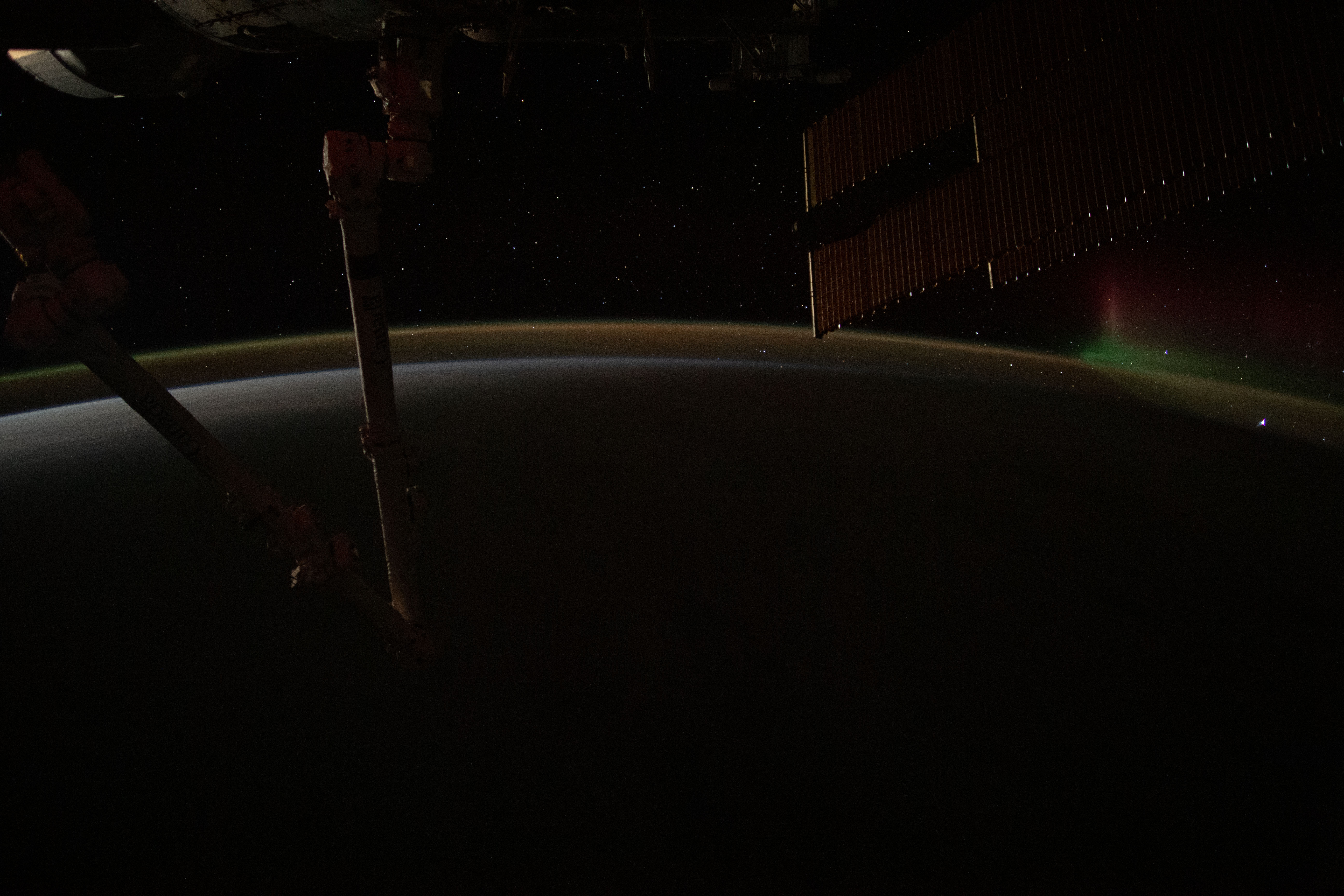 A wispy aurora fades away above Earth's atmospheric glow as the International Space Station orbits into a sunrise 271 miles above the Pacific Ocean southeast of New Zealand. In the foreground (from left) are, the Canadarm2 robotic arm and a pair of the orbiting lab's main solar arrays.