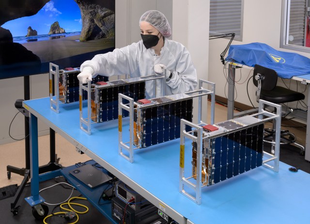A person wearing a lab coat, gloves, a hair net, and a face mask, grips a metal frame that supports a cereal box-sized small satellite, or CubeSat.
