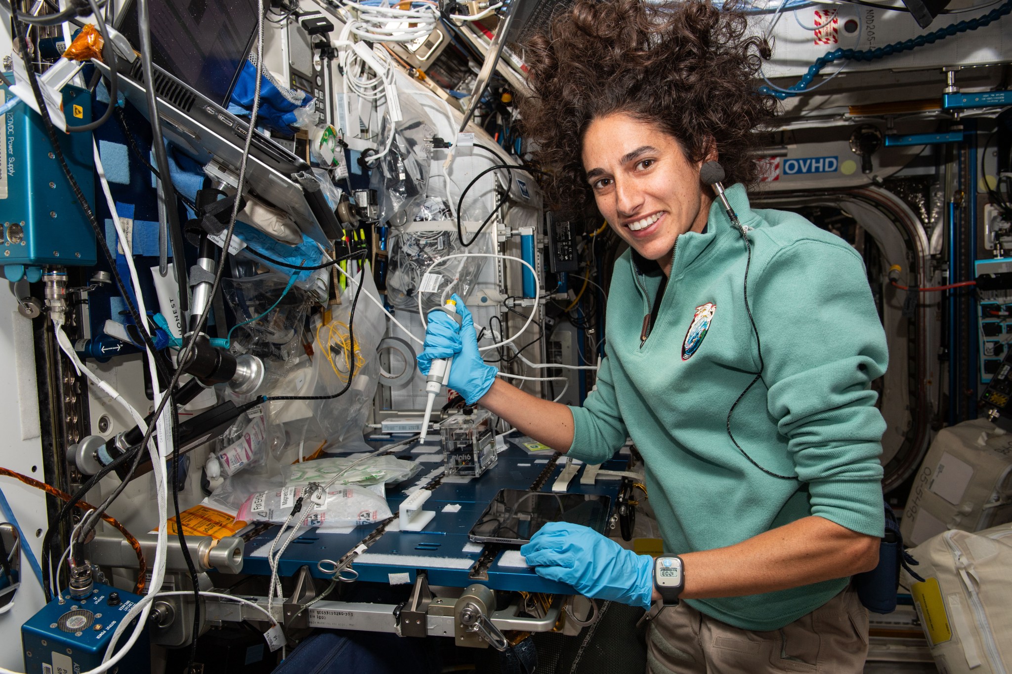 Jasmine Moghbeli holds a pipette used for water samples while working at a table. Her hair is floating around her head and a microphone floats by her ear.