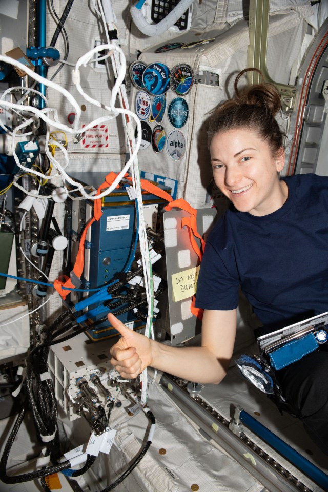 NASA astronaut and Expedition 67 Flight Engineer Kayla Barron begins her work day inside the International Space Station's Columbus laboratory module. She gives a "thumbs up" and poses next to the Light Ions Detector that monitors the radiation environment aboard the orbiting lab.