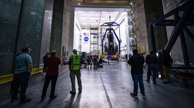 A scattered group of technicians and engineers stand inside the Space Environments Complex at NASA’s Neil Armstrong Test Facility. The group watches as the Dream Chaser spacecraft is moved into position for testing via a large crane.