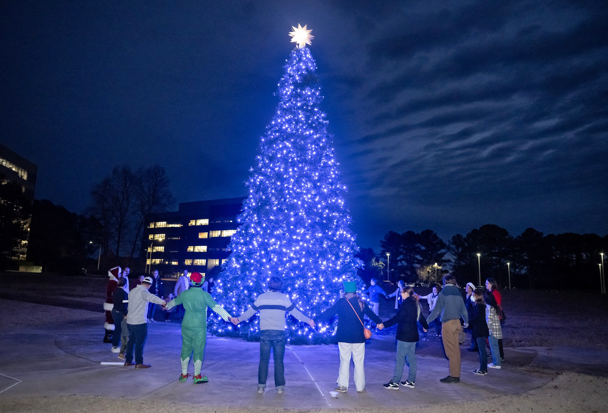 NASA Marshall Space Flight Center team members and family members form a circle as they bask in the light of the 32-foot artificial tree decorated with blue lights and a ten-pointed star representing each NASA center.