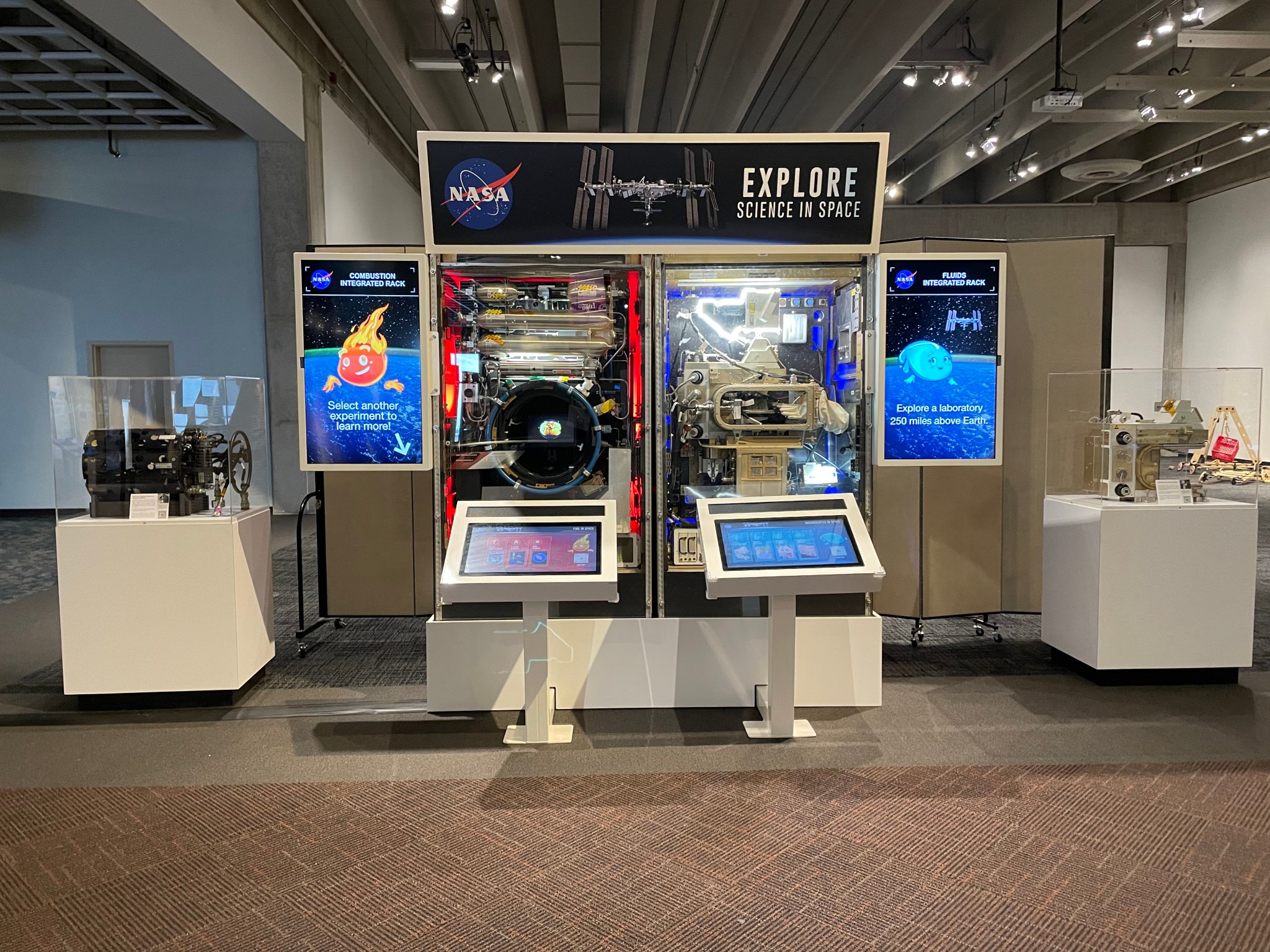 An exhibit with two touch-screen monitors and colorful video screens that feature animations and read “Combustion Integrated Rack” and “Fluids Integrated Rack” on either side. Text on a sign at the top of the exhibit reads, “Explore Science in Space.” A NASA meatball logo and an image of the International Space Station are also on the sign. Real space station hardware can be seen between the video monitors and on either side of the exhibit.