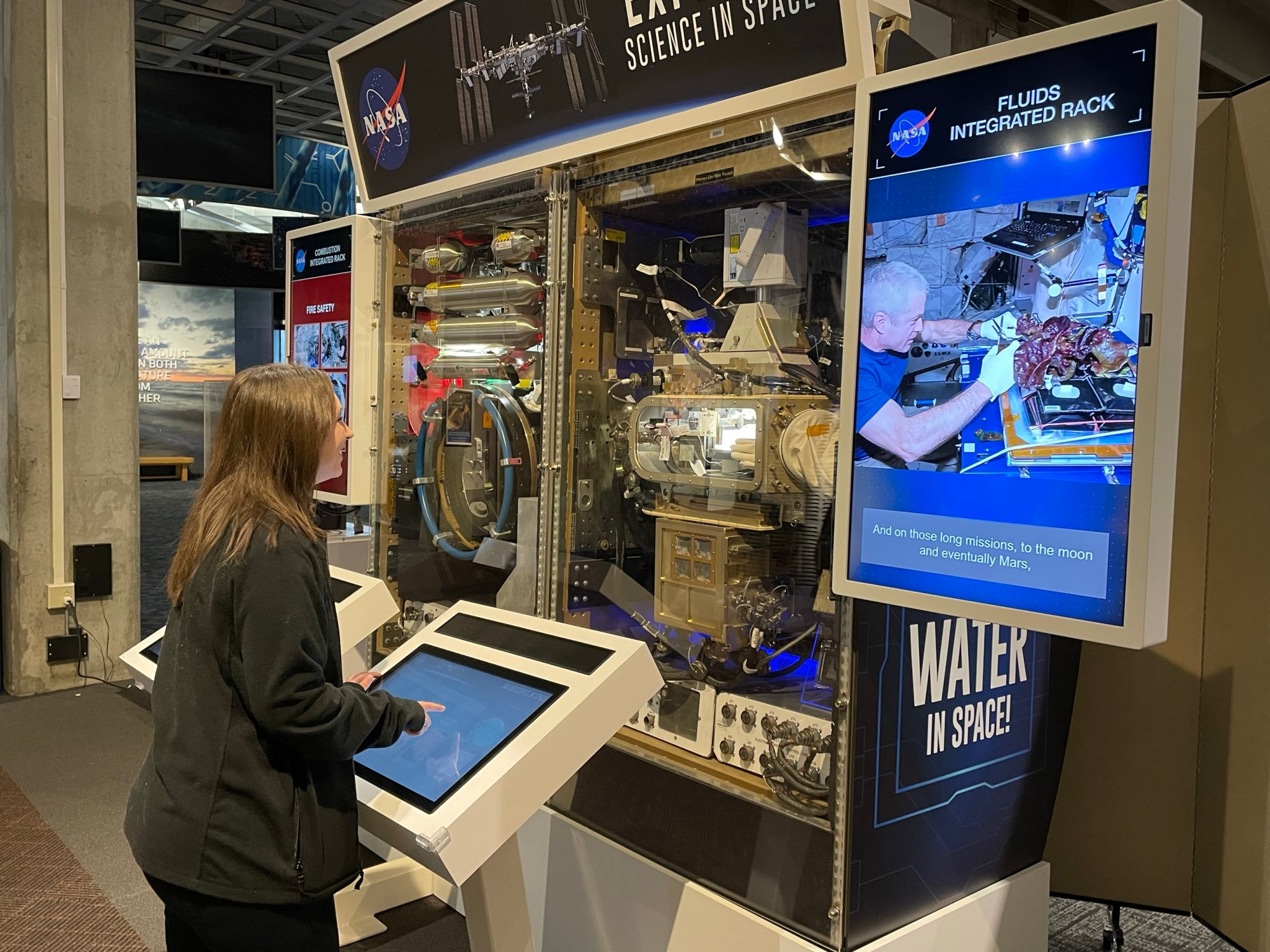 A woman visitor smiles as she operates an interactive space science exhibit using a colorful touch screen. Real International Space Station hardware can be seen between two video monitors in front of her and next to the exhibit.