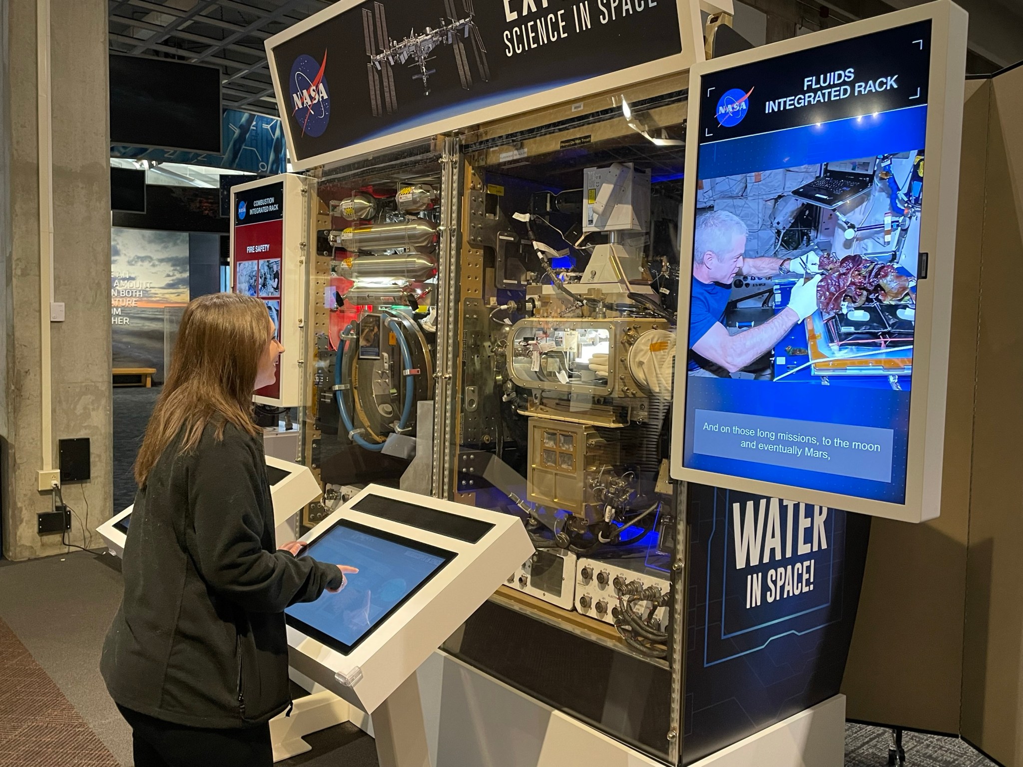 A woman visitor smiles as she operates an interactive space science exhibit using a colorful touch screen. Real International Space Station hardware can be seen between two video monitors in front of her and next to the exhibit.