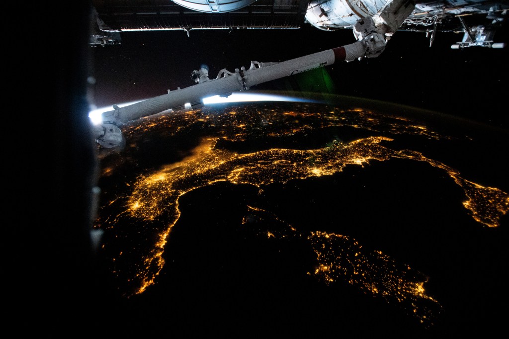 The city lights of Italy, including the French island of Corse and Italian islands of Sardinia and Sicily, are pictured from the International Space Station from 261 miles above the Mediterranean Sea while flying into an orbital sunrise. In the top foreground, is a portion of the Candarm2 robotic arm.