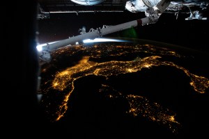 The city lights of Italy, including the French island of Corse and Italian islands of Sardinia and Sicily, are pictured from the International Space Station from 261 miles above the Mediterranean Sea while flying into an orbital sunrise. In the top foreground, is a portion of the Candarm2 robotic arm.