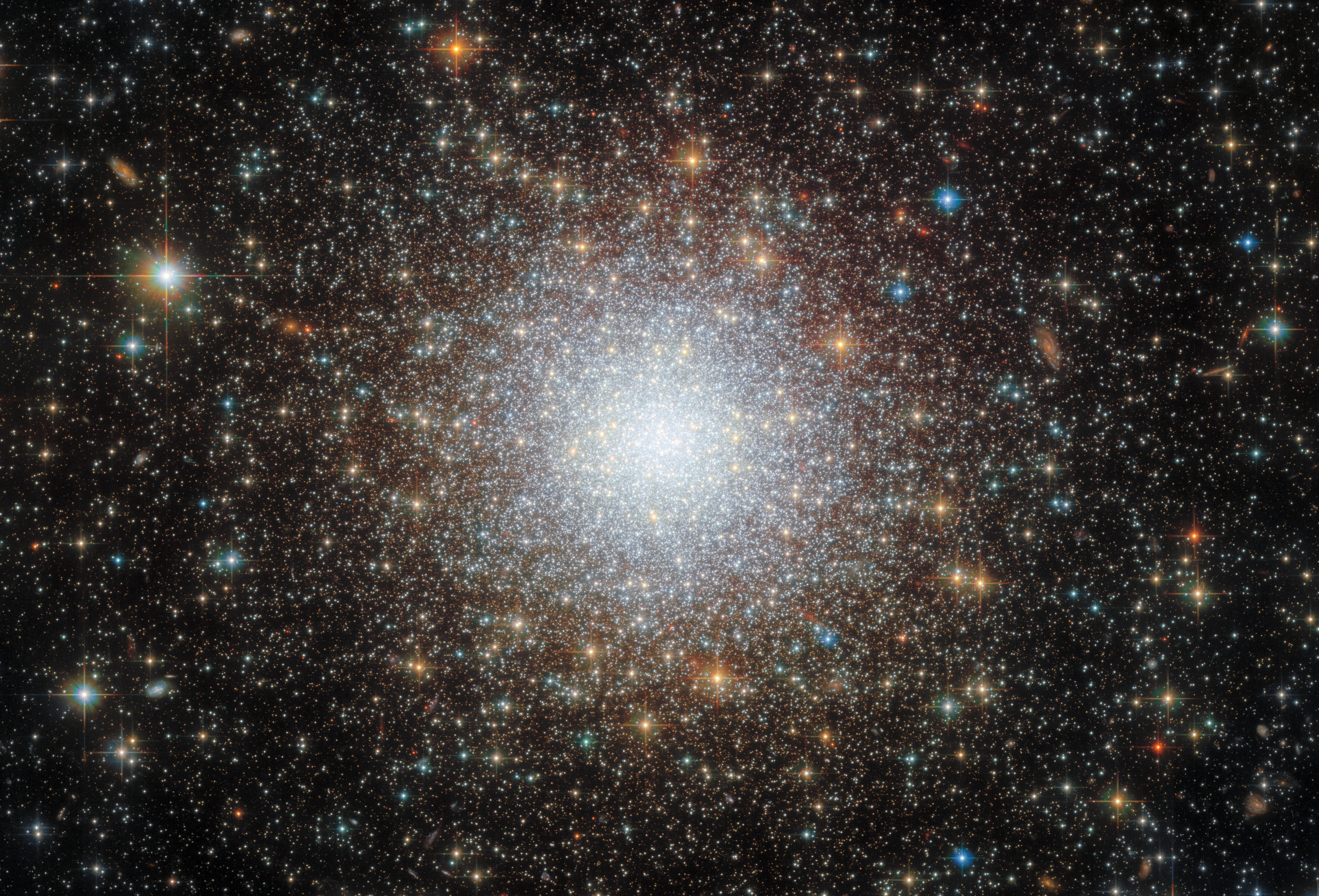 A dense cluster of stars. It is brightest and most crowded in the center, where the stars are mostly a cool white color. Moving out towards the edges the stars become more spread out and reddish until a noticeable ‘edge’ to the cluster is reached. Beyond that edge there are still many stars, more disorganized and seen on a black background. Some stars appear to be in front of the cluster.